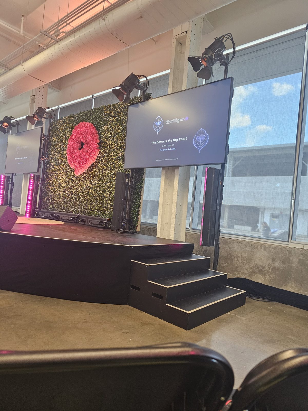The Village Stage at Startupfest 2026 — our title card on both screens: The Demo Is the Org Chart · distilligent.ai · The brain that asks.